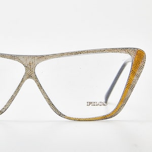 Könnte beinhalten: Katzenaugenbrille mit einem strukturierten beigen und grauen Schlangenledermuster auf dem Rahmen. Die Außenkanten weisen vertikale Akzente in einem leuchtenden Senfgelb auf. Das rechte Glas trägt den Schriftzug FILOS in Serifenschrift.