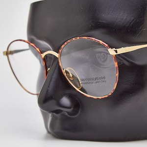 May include: Round eyeglasses with gold rims and tortoise shell frames. The glasses are displayed on a black mannequin head.