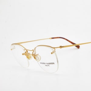 May include: Rimless spectacles with a thin gold-tone metal frame and bridge. The clear lenses feature the text Augusto Valentini Gold Pl printed on the right side. The temples are slender gold metal with brown tortoiseshell-patterned tips.
