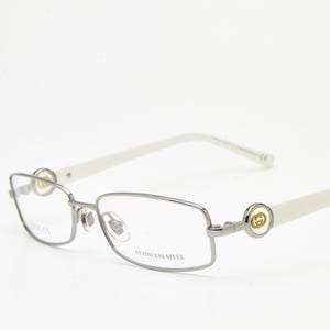 May include: Rectangular spectacles featuring a metallic silver-tone stainless steel frame and off-white temples. The hinges are adorned with a circular gold-tone logo emblem. The right lens is etched with the word Gucci and the left with Stainless Steel.