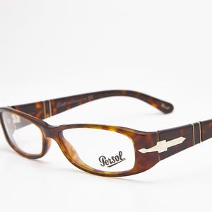 May include: Tortoiseshell spectacles featuring a brown and amber mottled pattern with a rectangular frame shape. The temples display a silver-toned arrow emblem near the hinges, and the right lens is printed with the brand name Persol in black.