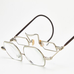 May include: Silver-toned metal spectacles featuring a unique double-lens design with octagonal frames. The glasses include thin, dark brown curved temple arms and small, light-coloured nose pads, creating a distinct, structured appearance.