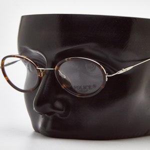 May include: Oval-shaped eyeglasses with a brown tortoiseshell pattern on the rims and thin silver-toned metal temples. The right lens features the word POLICE printed in a dark font. The glasses are displayed on a dark, abstract head form.