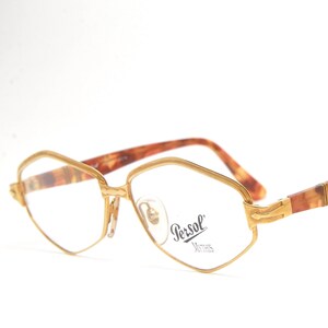 May include: Gold-toned metal eyeglasses with a geometric frame shape and brown tortoiseshell patterned temples. The right lens features the Persol brand logo and the word Mythos printed in black. The glasses have adjustable nose pads.