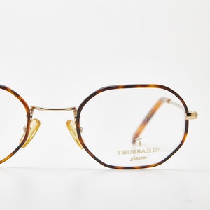 May include: Tortoiseshell-patterned eyeglasses with a gold-tone metal frame and bridge. The lenses are hexagonal in shape. The right lens features the text TRUSSARDI glasses with a small logo. The nose pads are gold-tone metal.