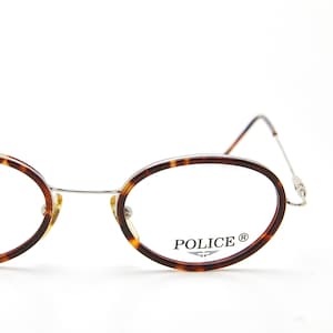May include: Oval-shaped spectacles featuring a brown and black tortoiseshell pattern on the rims, paired with thin silver metal temples and a bridge. The right lens displays the brand name POLICE with a small wing logo printed in black ink.