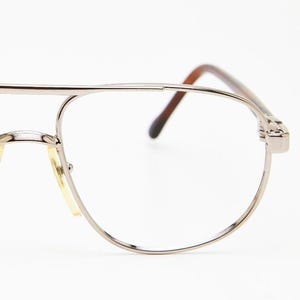 May include: Silver-toned aviator eyeglasses with clear lenses. The frames have a double bridge and brown temples. The nose pads are a light yellow color. The glasses are set against a white background.