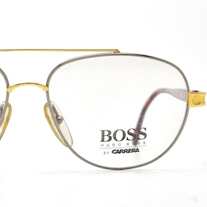 May include: Close-up of BOSS by Carrera spectacles. The spectacles have a gold-coloured top bar and accents, with a silver-coloured frame around the lenses. The lenses are clear, and the BOSS logo is visible on the right lens.