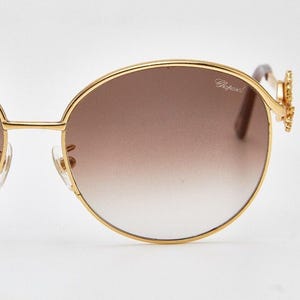 Könnte beinhalten: Goldrand-Sonnenbrille mit braunen Farbverlaufsgläsern. Das Chopard-Logo ist auf dem Glas zu sehen. Die Bügel haben ein dekoratives Blumenmuster. Ein modisches Accessoire zum Schutz der Augen.