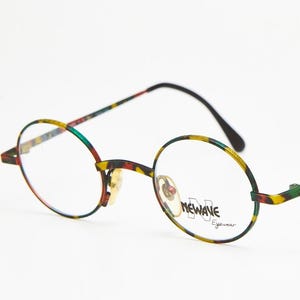 Può includere: Occhiali rotondi con un motivo colorato sulle montature e sulle aste. Le montature sono nere, con un motivo giallo, verde e rosso. Le lenti sono trasparenti e il marchio "NEWAVE Eyewear" è visibile.