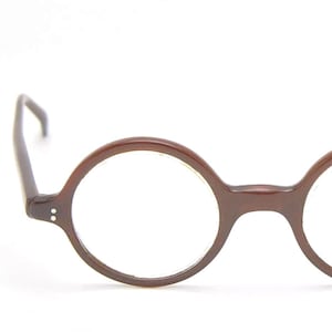 May include: A pair of round-framed spectacles with a dark brown, polished finish. The frames feature a circular lens shape, a central bridge, and two small silver-toned rivets on each outer corner where the temples attach to the front frame.