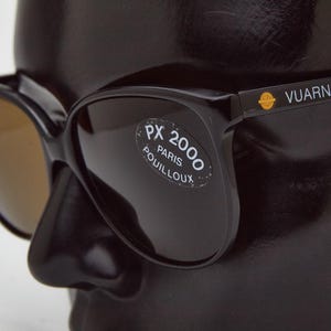 May include: Black cat-eye sunglasses with brown lenses. The sunglasses have a gold label that reads "VUARNET PX 2000 PARIS POUILLOUX".