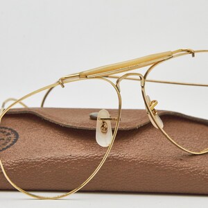 May include: Gold-tone metal aviator glasses featuring a prominent brow bar with a light-colored plastic insert and clear lenses. The glasses are positioned in front of a textured brown leather-like case with a visible Ray-Ban logo stamp.