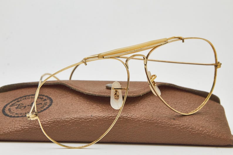 May include: Gold-tone metal aviator glasses featuring a prominent brow bar with a light-colored plastic insert and clear lenses. The glasses are positioned in front of a textured brown leather-like case with a visible Ray-Ban logo stamp.