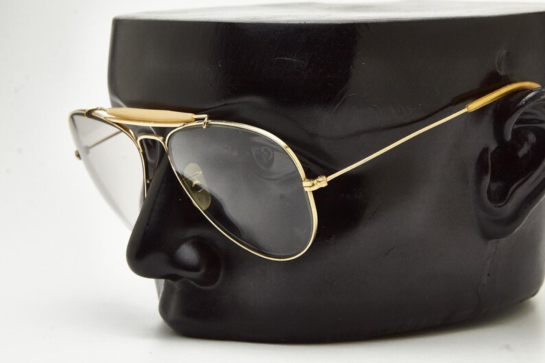 Puede incluir: Gafas de sol de aviador con montura dorada y lentes oscuras. Las gafas se exhiben en una cabeza de maniqu&iacute; negra, mostrando el dise&ntilde;o y la forma de las gafas. Las gafas tienen un puente y patillas doradas.