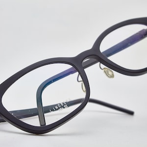 May include: Black rectangular eyeglasses with a thin metal frame and clear lenses. The brand name "LINDBERG" is printed on the right temple.