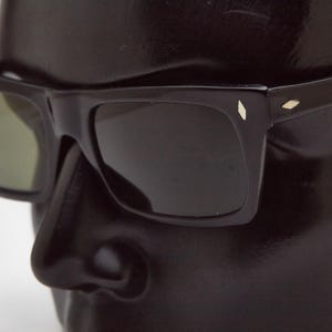 May include: Black sunglasses with a rectangular frame and green-tinted lenses. The sunglasses have a small, decorative diamond-shaped accent on each temple. The sunglasses are displayed on a black mannequin head.