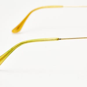 Puede incluir: Primer plano de gafas con montura de alambre dorado y patillas amarillo-verdosas. Las patillas se curvan suavemente, terminando en una forma redondeada. El fondo es blanco liso.