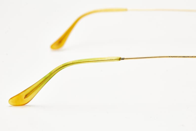 Puede incluir: Primer plano de gafas con montura de alambre dorado y patillas amarillo-verdosas. Las patillas se curvan suavemente, terminando en una forma redondeada. El fondo es blanco liso.