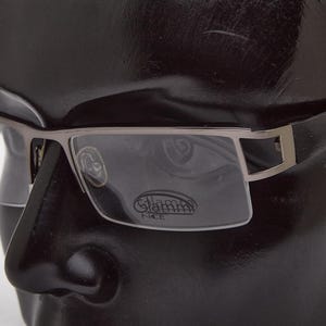 May include: Eyeglasses with a rectangular frame and a silver-colored metal bridge. The lenses are clear, and the temples are dark. The brand name "Glamm Nice" is visible on the lens.