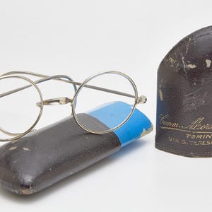 Może przedstawiać: Okulary vintage z okrągłą oprawką w kolorze srebrnym leżą na czarno-niebieskim etui. Drugie, pasujące etui stoi pionowo. Etui jest ciemnobrązowe ze złotym napisem "Comm. Alborochin, TORINO, VIA S. TERESA O."