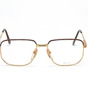 May include: Gold-tone eyeglasses with a rectangular frame and brown accents. The glasses have a classic design with a double bridge and nose pads. The temples are thin and extend to the ear pieces. The lenses are clear.