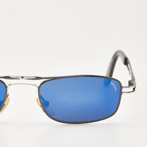 May include: Daytona sunglasses with a black frame and blue lenses. The sunglasses have a rectangular shape and a silver bridge. The text "DAYTONA" is visible on the right lens.