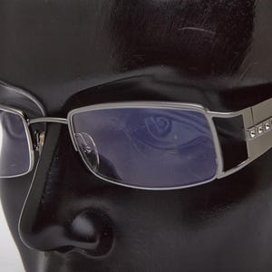 May include: Rectangular eyeglasses with a silver frame and clear lenses. The temples are a darker silver with small, decorative rhinestones. The glasses are displayed on a black mannequin head.