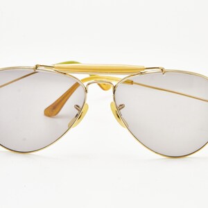 Puede incluir: Gafas de sol aviador con montura dorada y lentes gris claro. Las gafas de sol tienen un doble puente y una barra superior, con detalles marrones y amarillos en las patillas y almohadillas nasales. Un accesorio de gafas cl&aacute;sico.