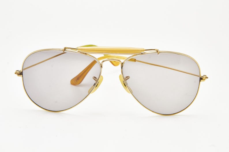 Puede incluir: Gafas de sol aviador con montura dorada y lentes gris claro. Las gafas de sol tienen un doble puente y una barra superior, con detalles marrones y amarillos en las patillas y almohadillas nasales. Un accesorio de gafas cl&aacute;sico.