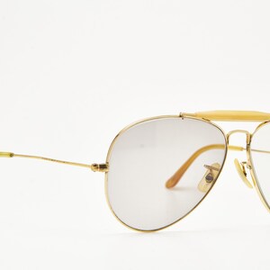 Puede incluir: Gafas de sol de aviador con montura dorada y lentes degradadas. Las gafas tienen un doble puente y una barra de color claro en la parte superior. Las patillas son doradas con puntas amarillas. Las lentes tienen un tinte degradado.