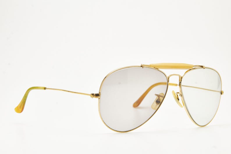 Puede incluir: Gafas de sol de aviador con montura dorada y lentes degradadas. Las gafas tienen un doble puente y una barra de color claro en la parte superior. Las patillas son doradas con puntas amarillas. Las lentes tienen un tinte degradado.