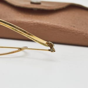 May include: A pair of gold-toned eyeglasses featuring a thin metal frame, a curved bridge, and a textured wire detail along the lower rim. The temple arm displays a small engraved marking near the hinge, and a brown protective case is visible.