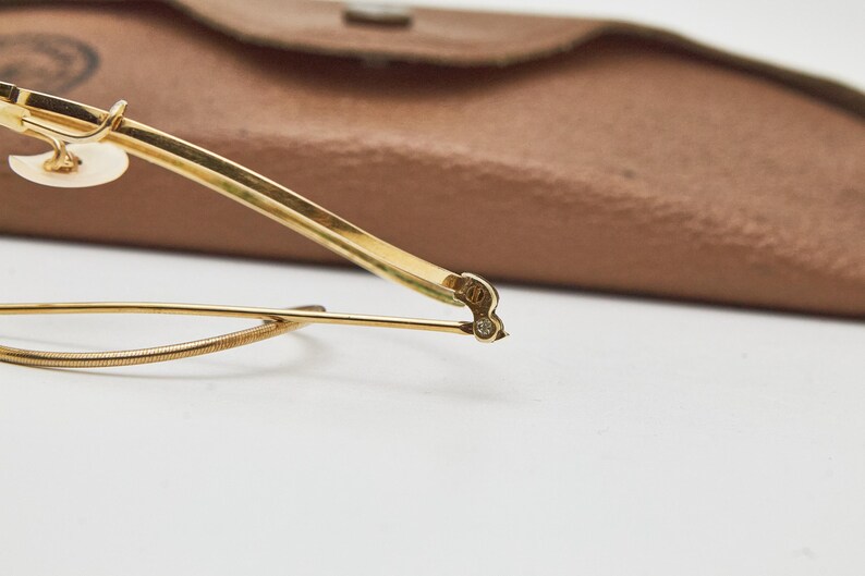 May include: A pair of gold-toned eyeglasses featuring a thin metal frame, a curved bridge, and a textured wire detail along the lower rim. The temple arm displays a small engraved marking near the hinge, and a brown protective case is visible.