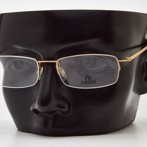 May include: Gold-rimmed eyeglasses with clear lenses on a black mannequin head. The brand name "Rodenstock" is visible on the right lens.