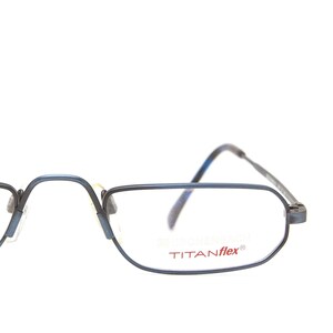 Può includere: Occhiali rettangolari blu con la scritta "TITANflex®" sulla lente. Le montature sono di colore blu scuro e hanno un design sottile, simile a un filo. Gli occhiali sono su uno sfondo bianco.
