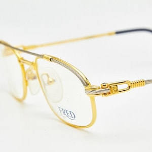 Pode incluir: Óculos de metal em tom dourado com design de ponte dupla e detalhes de fio trançado em tom prateado ao longo da borda superior e das hastes. As lentes transparentes têm a palavra FRED impressa em azul e as pontas das hastes são pretas.