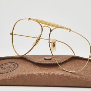May include: Gold-tone metal aviator glasses with a prominent brow bar and clear lenses, resting on a brown textured leather-like case featuring a circular Ray-Ban logo stamp with the text "Ray-Ban" and "100% UV PROTECTION" printed in black.
