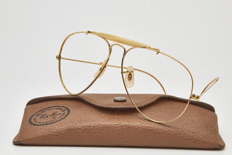 May include: Gold-tone metal aviator glasses with a prominent brow bar and clear lenses, resting on a brown textured leather-like case featuring a circular Ray-Ban logo stamp with the text "Ray-Ban" and "100% UV PROTECTION" printed in black.