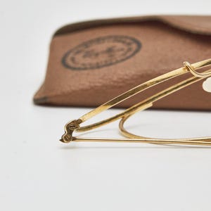 May include: Gold-tone metal eyeglasses featuring thin wire frames, a curved bridge, and clear nose pads. The glasses are positioned in front of a tan, textured leather-like carrying case with a circular logo stamp visible on the surface.