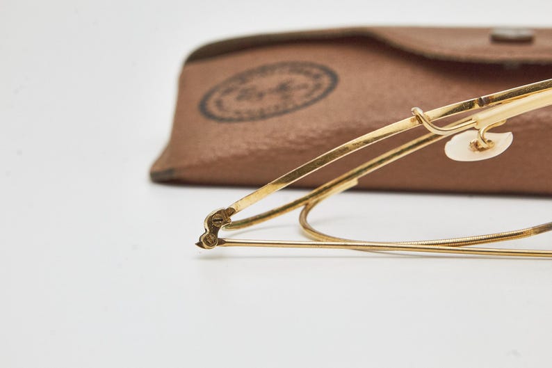 May include: Gold-tone metal eyeglasses featuring thin wire frames, a curved bridge, and clear nose pads. The glasses are positioned in front of a tan, textured leather-like carrying case with a circular logo stamp visible on the surface.