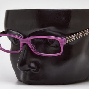 Könnte beinhalten: Eine Brille mit lila Rahmen und rechteckigen Gläsern liegt auf einem schwarzen, kopfartigen Display. Die Bügel der Brille sind silberfarben und tragen den Schriftzug "MARC BY MARC JACOBS". Die Brille hat eine transparente lila Farbe.
