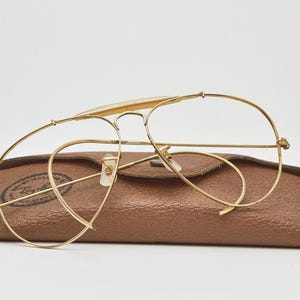 May include: Gold-tone metal aviator glasses with thin wire frames and clear lenses, resting on a textured brown leather-like protective case. The case features a circular logo with the text Ray-Ban printed in black on the left side.