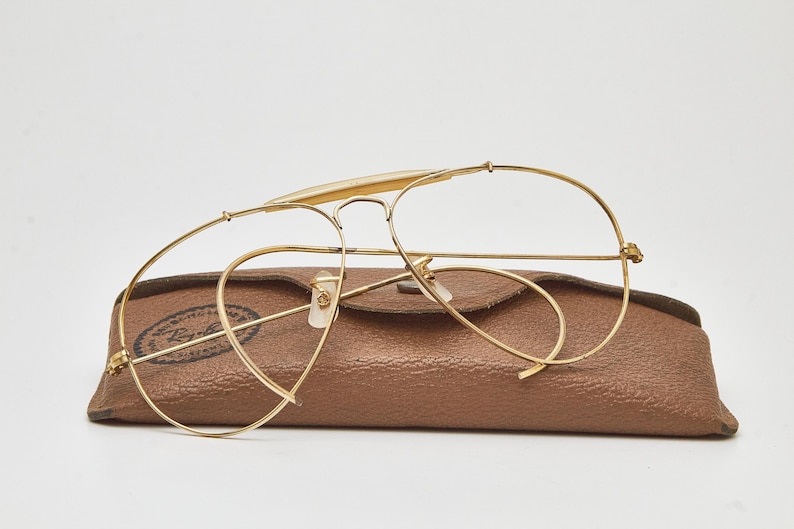 May include: Gold-tone metal aviator glasses with thin wire frames and clear lenses, resting on a textured brown leather-like protective case. The case features a circular logo with the text Ray-Ban printed in black on the left side.