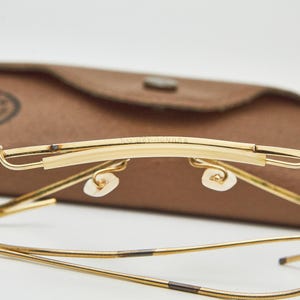 May include: Gold-tone metal eyeglasses featuring a thin wire frame and a brow bar with the text B&L RAY-BAN U.S.A. engraved on the top. The glasses include adjustable nose pads and flexible cable temples, presented with a brown textured case.