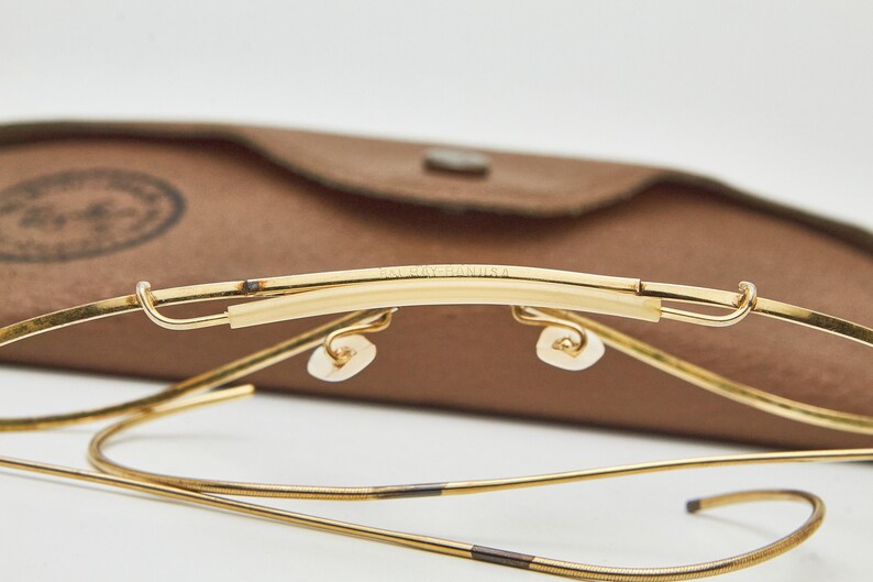 May include: Gold-tone metal eyeglasses featuring a thin wire frame and a brow bar with the text B&L RAY-BAN U.S.A. engraved on the top. The glasses include adjustable nose pads and flexible cable temples, presented with a brown textured case.