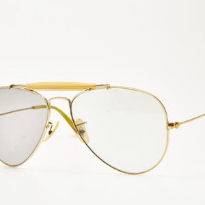 Puede incluir: Gafas de sol de aviador con montura dorada, lentes transparentes y puente de color claro. Las patillas tienen un dise&ntilde;o curvo con un color marr&oacute;n claro. Las lentes son de color gris plateado.