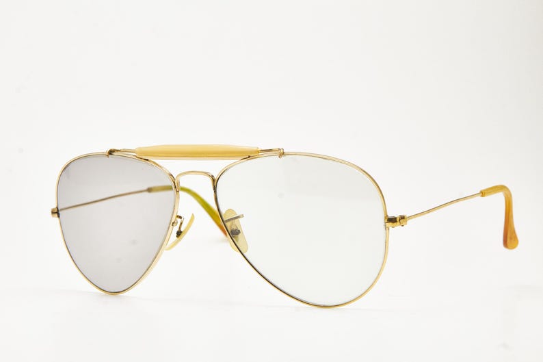 Puede incluir: Gafas de sol de aviador con montura dorada, lentes transparentes y puente de color claro. Las patillas tienen un dise&ntilde;o curvo con un color marr&oacute;n claro. Las lentes son de color gris plateado.