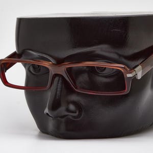 May include: Brown rectangular eyeglasses with a reddish-brown frame are displayed on a black mannequin head. The glasses have a modern design with a silver accent on the temples. The mannequin head is a dark, glossy color.