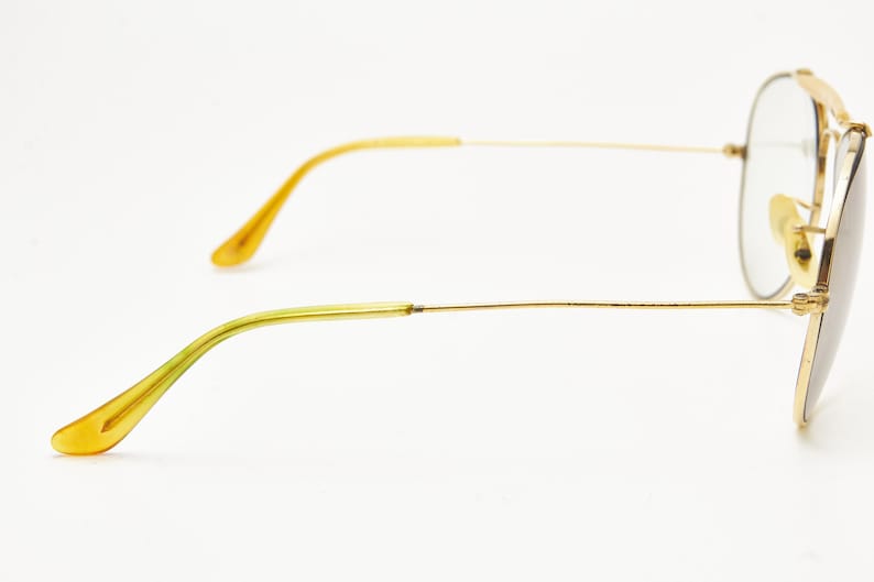 Puede incluir: Gafas de sol de aviador con montura dorada y lentes transparentes. Las patillas son finas y doradas, con una transici&oacute;n a un tono amarillo-verdoso en los terminales. Las lentes son de color gris claro y las almohadillas nasales son doradas.